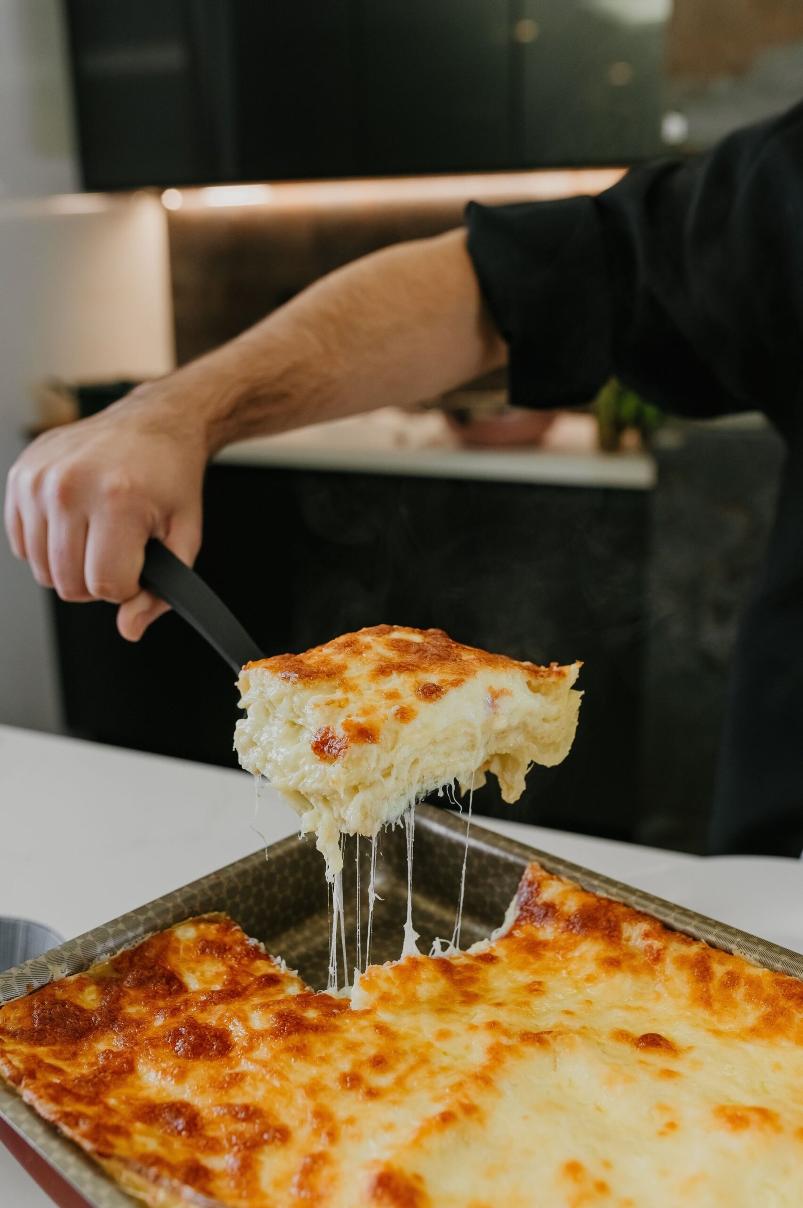 Close-up of a gooey, cheesy slice of lasagna being served from a baking tray in a cozy kitchen setting.